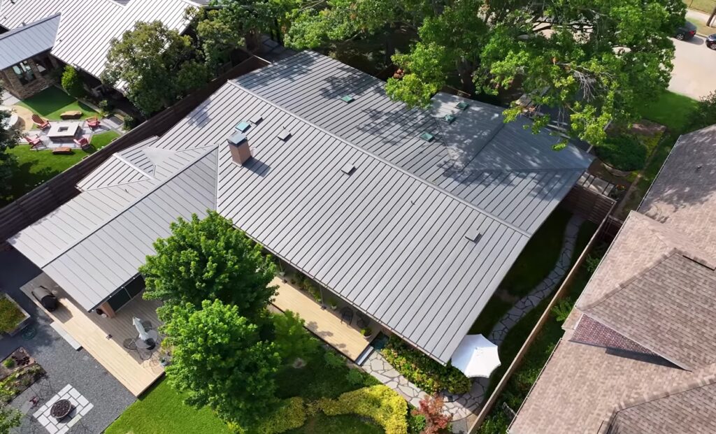 High-angle drone photograph of a modern residential home showcasing a newly installed dark grey standing seam metal roof. The property features intricate roof valleys, a chimney, skylights, beautifully landscaped yards, a stone pathway, and a wooden deck.