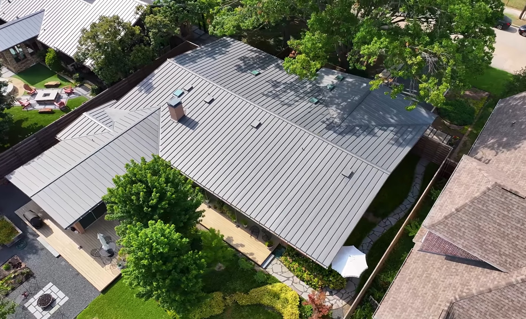 High-angle drone photograph of a modern residential home showcasing a newly installed dark grey standing seam metal roof. The property features intricate roof valleys, a chimney, skylights, beautifully landscaped yards, a stone pathway, and a wooden deck.