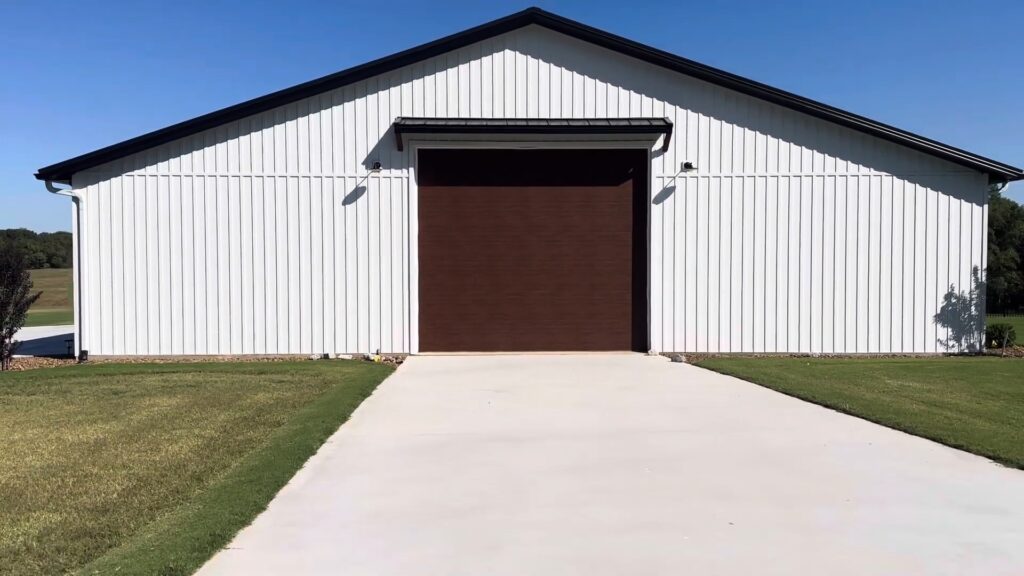 A large modern pole barn building featuring white vertical board and batten metal siding and a dark roof trim. A concrete driveway leads up to a wide, dark brown wood-look roll-up garage door centered on the front facade under a clear blue sky.