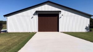 A large modern pole barn building featuring white vertical board and batten metal siding and a dark roof trim. A concrete driveway leads up to a wide, dark brown wood-look roll-up garage door centered on the front facade under a clear blue sky.
