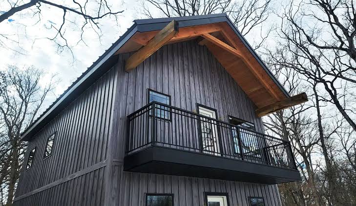 A photorealistic exterior shot of a striking modern farmhouse featuring matte black metal board and batten siding. The vertical lines of the siding are prominent and seamless. The home features warm wood accents under the eaves, large glass windows, and a sleek modern aesthetic.